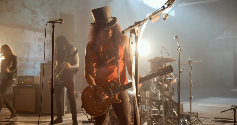 Image of Slash Playing Guitar on Stage with Myles Kennedy and The Conspirators, Wearing a Top Hat and A Red Sleeveless Shirt, Under Dramatic Stage Lighting